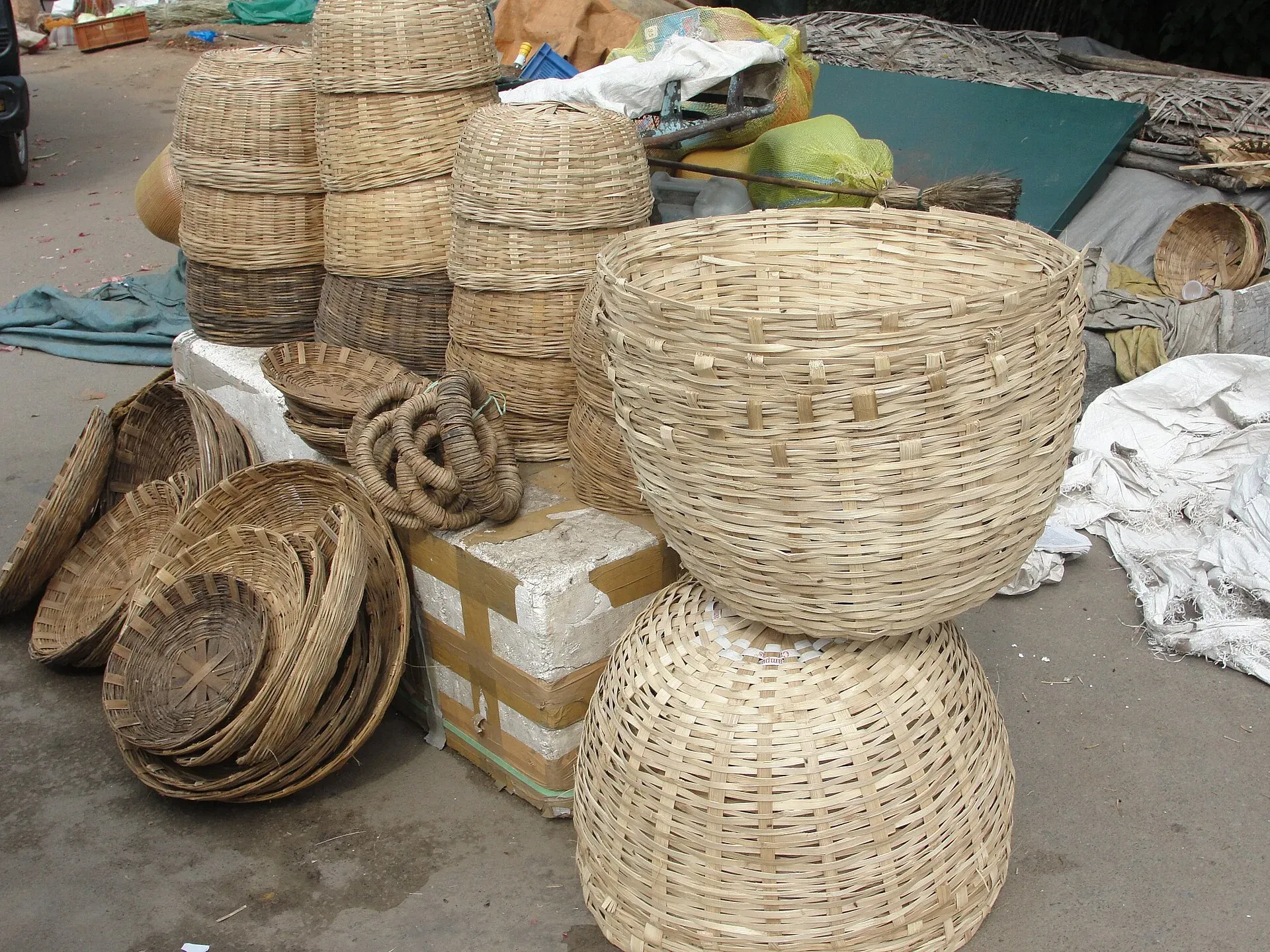 Bamboo and cane baskets stacked at a roadside market — lidded storage baskets, shallow winnowing trays, coils of woven cane and two large open-weave baskets in the foreground