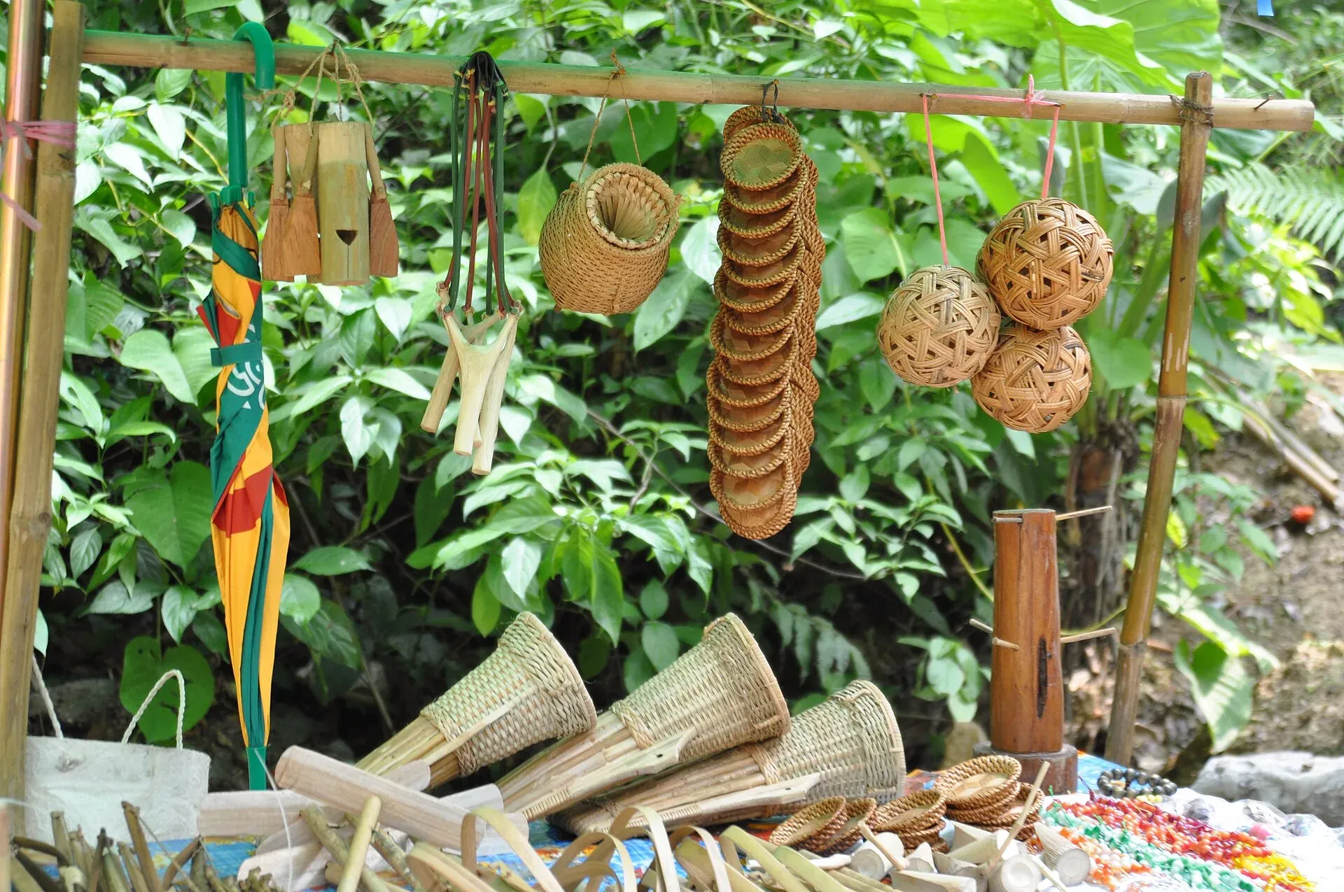 A roadside craft stall against green forest — woven cane balls, stacked coasters, a small basket, a bamboo mug and conical fish traps hung from and spread beneath a bamboo pole
