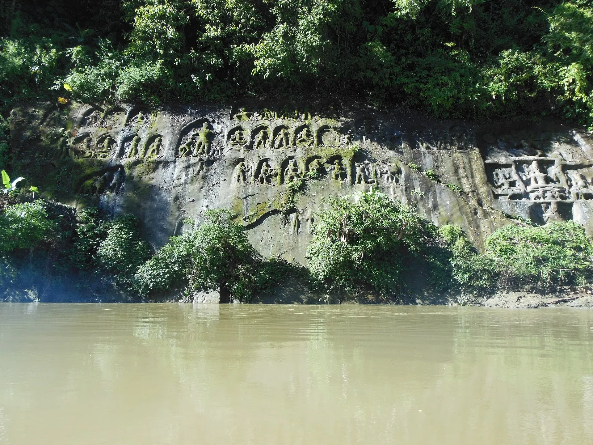 Rows of moss-tinged figures carved into the dark rock face at Chabimura, rising directly from the green water of the Gomati River beneath overhanging forest