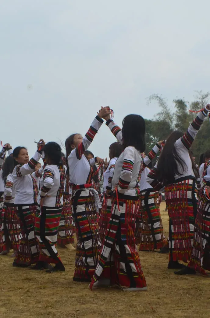 Dancers at Chapchar Kut in white tops with banded sleeves and striped red, black and white skirts, arms raised and hands joined overhead on an open festival ground