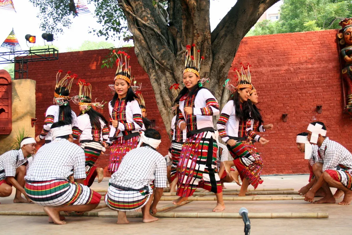 A Cheraw performance — women in headdresses and patterned skirts stepping between long bamboo staves clapped together by kneeling men in checked shirts, under a tree against a red brick wall
