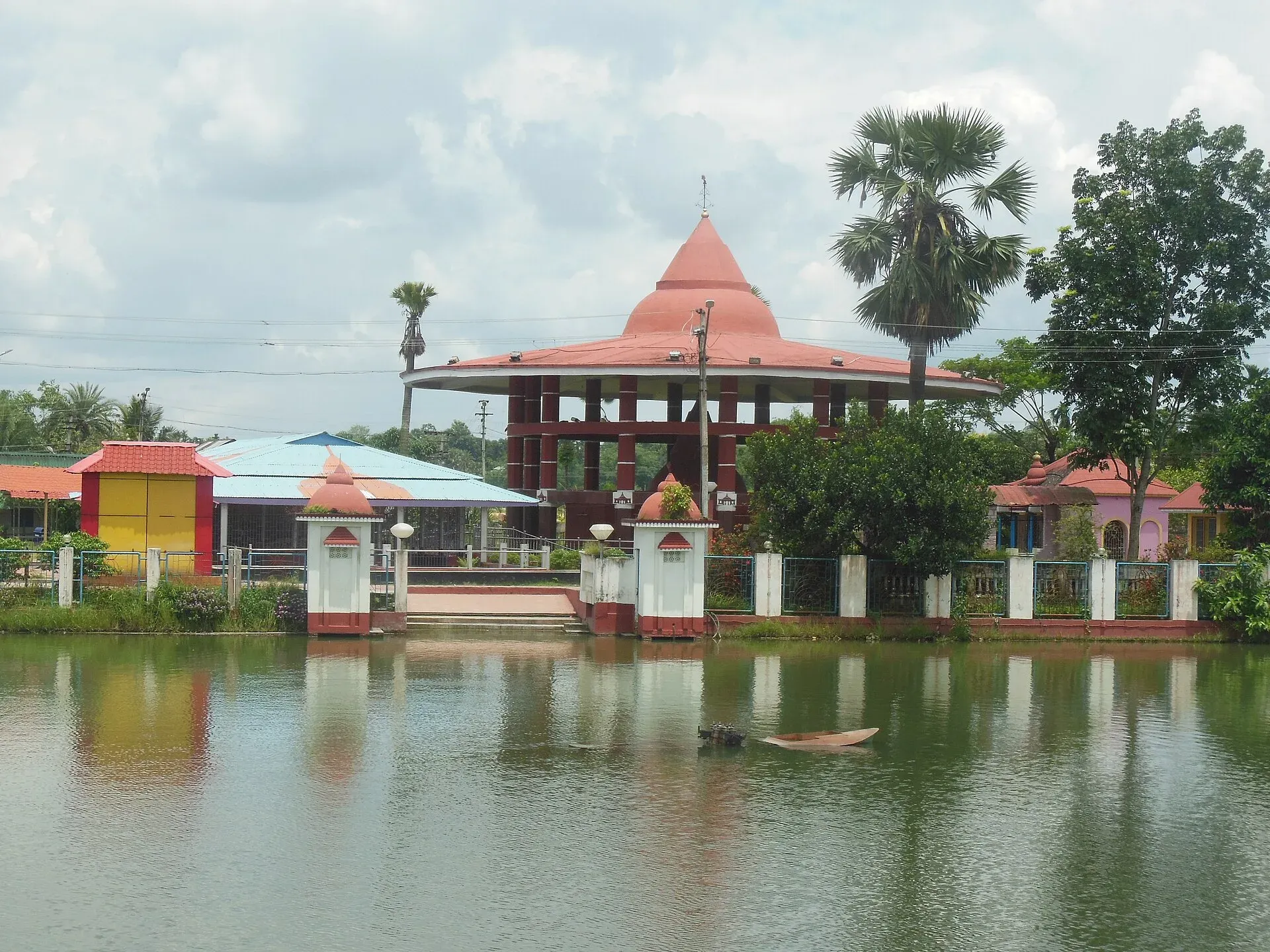 The temple of the Fourteen Deities at Old Agartala seen across its tank — a conical orange-roofed pavilion on dark red columns behind white gateposts, palms and a fenced ghat