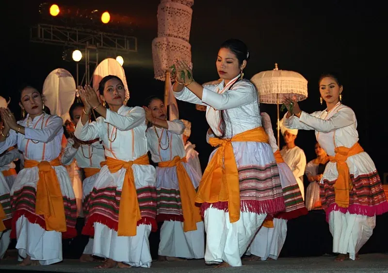 Maibi dancers at Lai Haraoba in white robes with orange sashes and striped red-and-green skirts, palms raised together in unison, a white ceremonial umbrella behind them