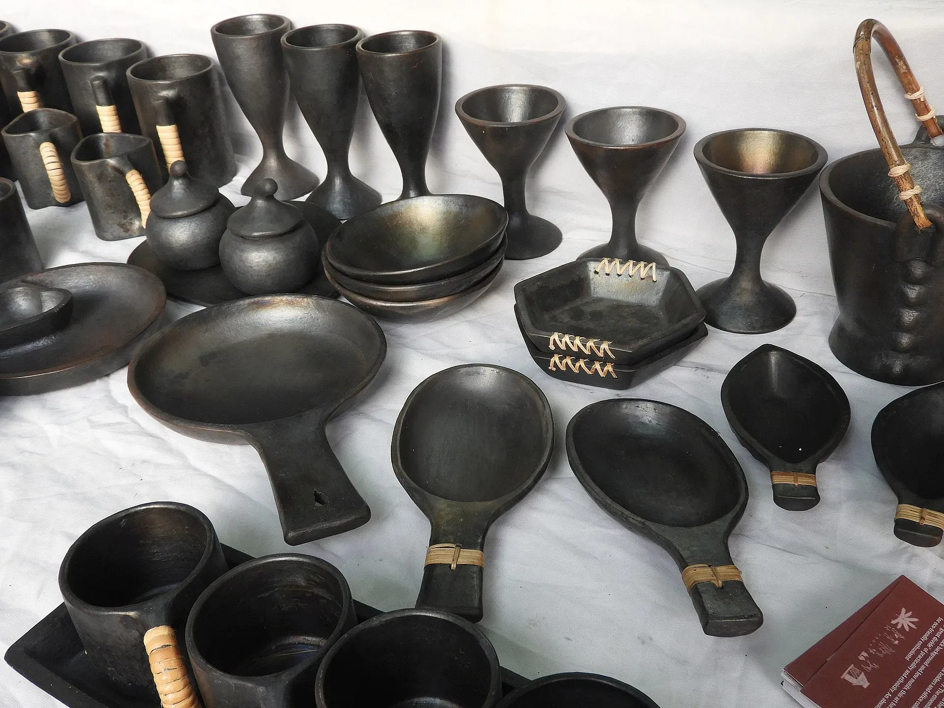 Longpi black pottery laid out on a white cloth — goblets, lidded pots, bowls, plates and spoon-shaped ladles in matte charcoal-black clay, several bound with woven cane