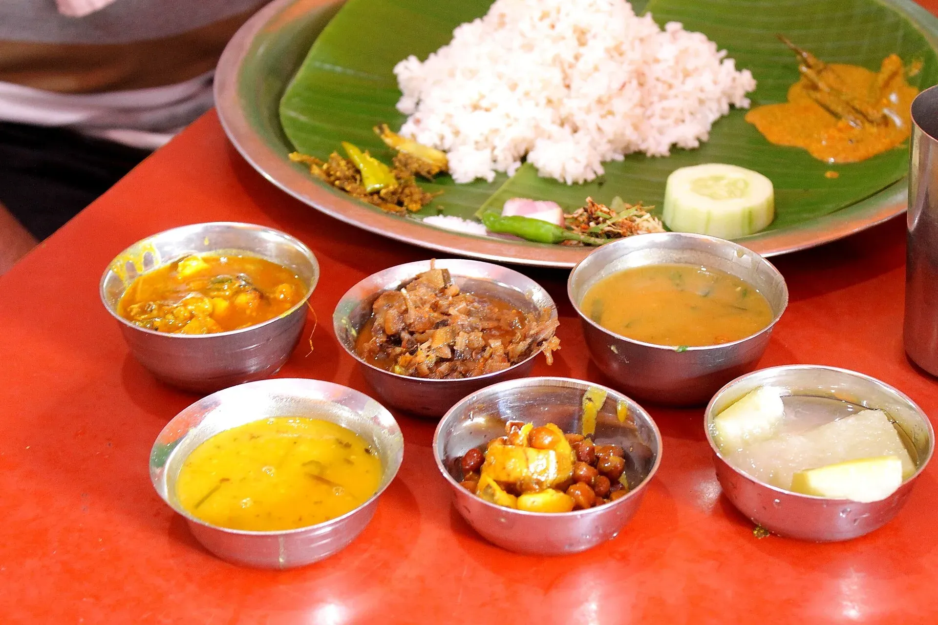 A Manipuri meal on a red table — rice with sides served on a banana leaf, ringed by steel bowls of dal, black chickpeas, chopped greens and fish curry