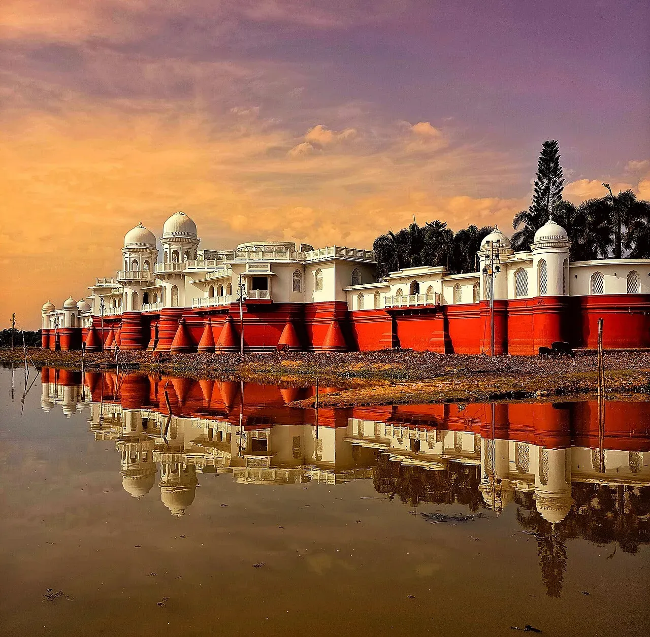 Neermahal palace on Rudrasagar Lake at sunset, white domes above red-banded walls mirrored in the still water