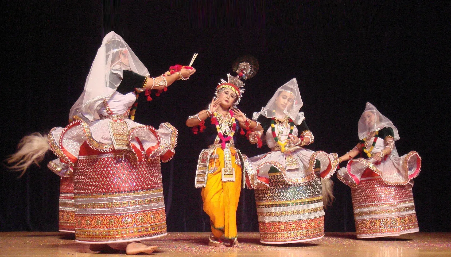 Ras Lila on stage — a dancer as Krishna in yellow with a peacock-feather crown at the centre, surrounded by dancers in stiff barrel-shaped red-and-silver skirts and translucent white veils