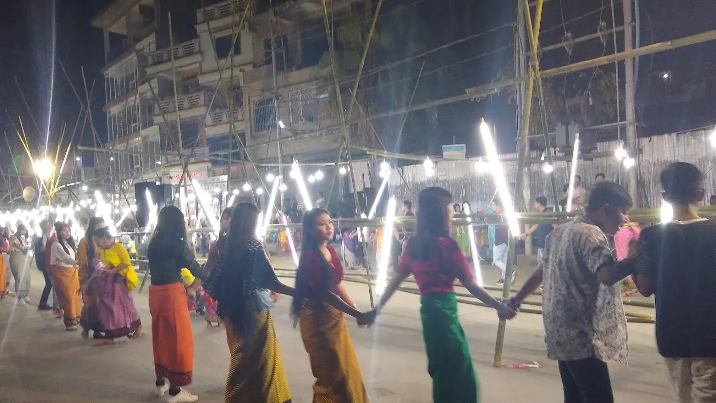 Thabal Chongba during Yaoshang in Imphal at night — dancers in striped sarongs holding hands in a long chain beneath strings of tube lights lashed to bamboo scaffolding