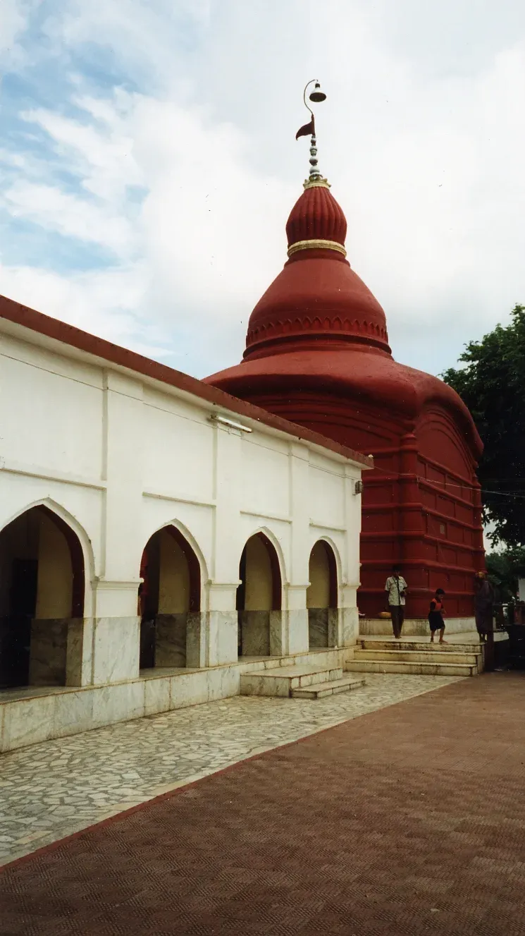 The Tripura Sundari Temple at Udaipur — a deep-red curved shikhara crowned with a finial rising behind the white arched pilgrim hall, visitors on the marble steps below