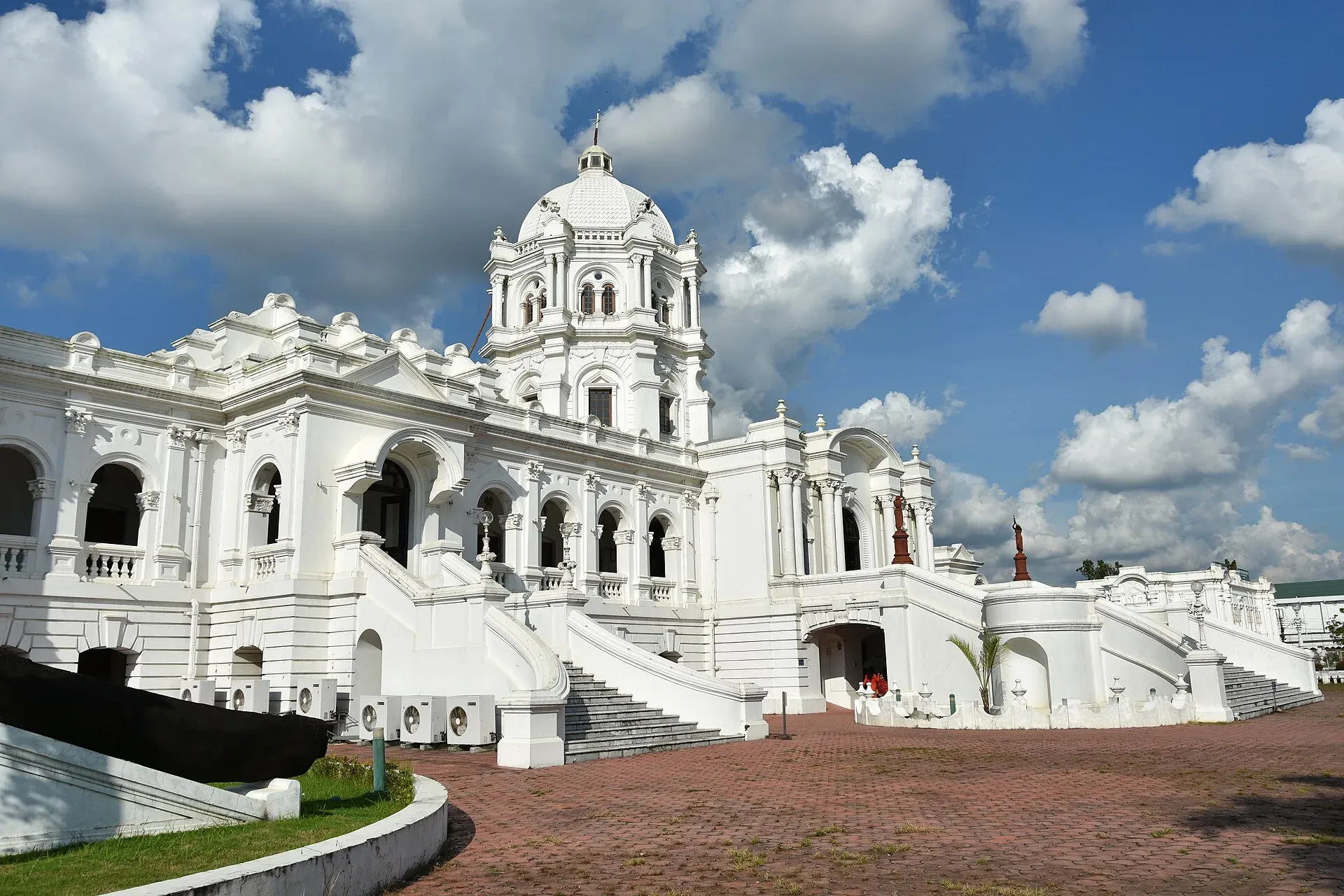 The white domed facade of Ujjayanta Palace in Agartala under a blue sky with drifting clouds, twin staircases curving up to the columned entrance from the brick forecourt