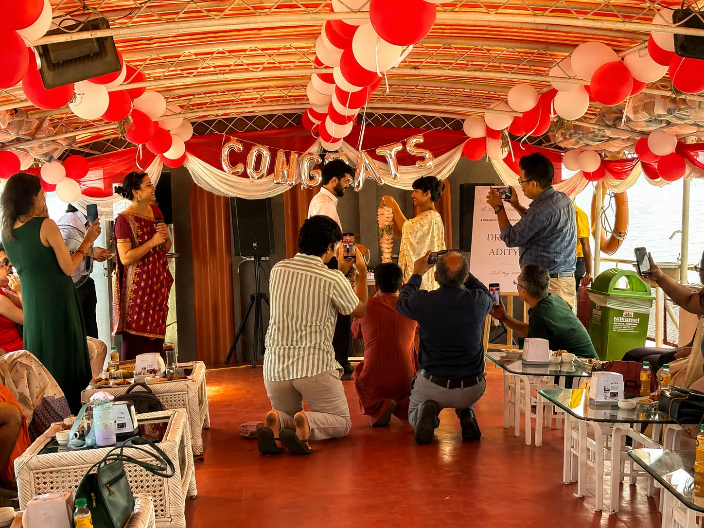 Couple exchanging garlands under balloons and a Congrats banner at an onboard celebration
